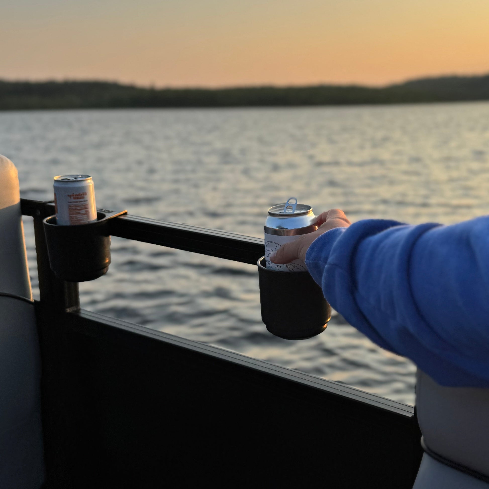 Person grabbing a can out of a cup holder on a pontoon boat with a sunset over water in the background